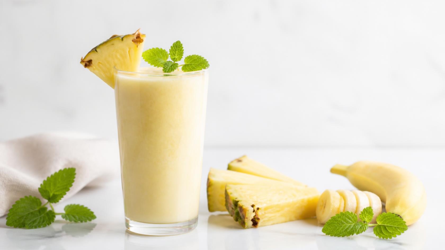 Ananászos smoothie hozzávalói letisztult elrendezésben: ananász, banán, citromfű, kókuszvíz és méz világos háttéren.